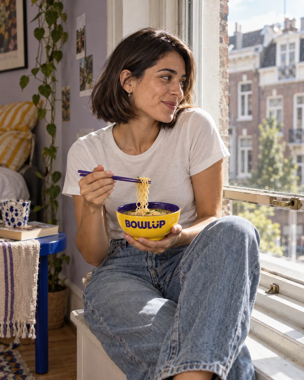A young woman takes a relaxed lunch break by a city apartment window, holding a yellow BOWLUP bowl and purple chopsticks.